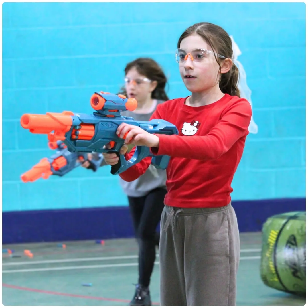 A girl in safety glasses aims a foam dart blaster in a sports hall, with a second child behind her