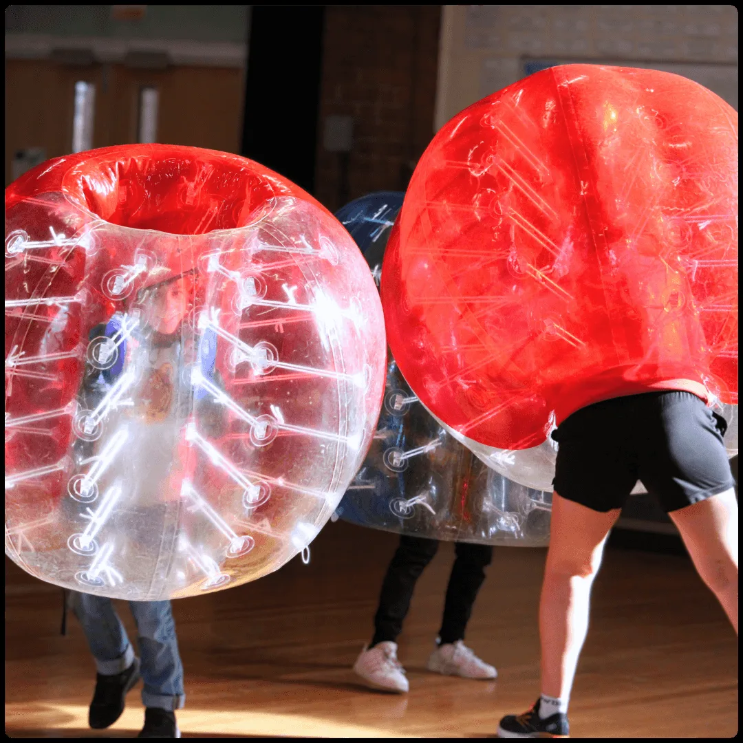 Children inside clear and red inflatable bubble football suits on a wooden sports hall floor